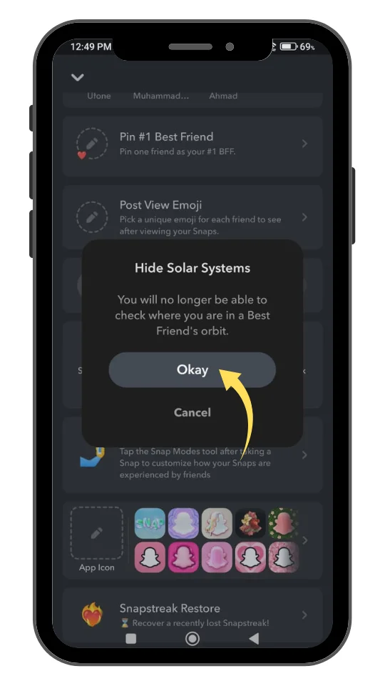 A smartphone screen showing a Snapchat settings confirmation popup. The popup message reads, "Hide Solar Systems — You will no longer be able to check where you are in a Best Friend's orbit." Below the message are two options: a highlighted "Okay" button with a yellow arrow pointing to it, and a "Cancel" button. The background screen displays Snapchat settings like "Pin #1 Best Friend," "Post View Emoji," and "Snapstreak Restore" in dark mode.