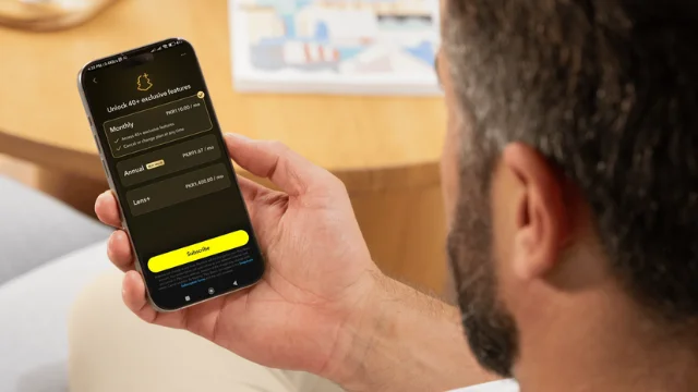 A man holding a smartphone displaying Snapchat+ subscription plans, including Monthly, Annual, and Lens options. The phone screen shows a yellow "Subscribe" button, and the background includes a blurred wooden table and magazine.